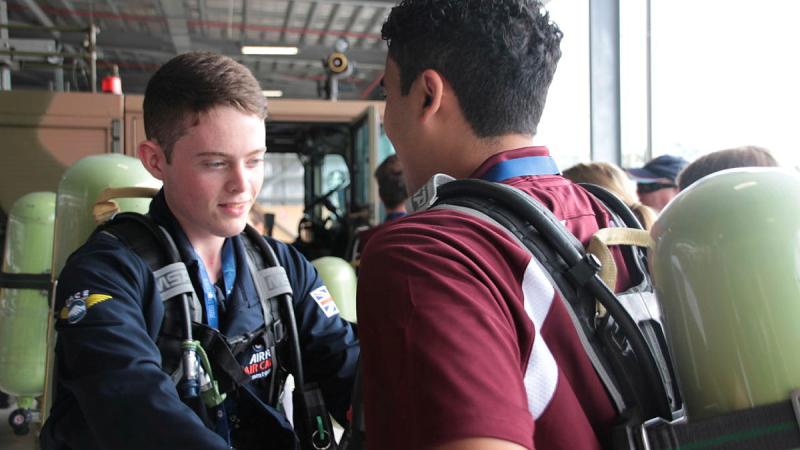 UK Air Cadet Alex Athey helps United States Air Cadet Ishan Swali into his breathing gear at an information session on flight line fire suppression at 23 Squadron during their visit. 