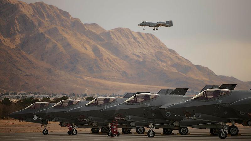 Royal Australian Air Force No. 77 Squadron F-35A Lightning II aircraft on the Nellis Air Force Base flight line during Exercise Red Flag Nellis 25-1 in the US, with Nevada hills in ther background and a US aircraft overhead..