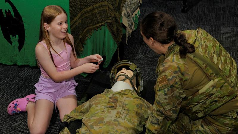 A girl in a pink outfit talks to a soldier in camouflage uniform about a dummy used to give health demonstrations.