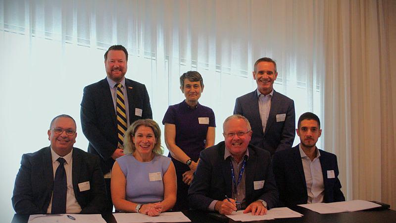 Representatives from Defence Science and Technology Group (DSTG), the University of South Australia and the Alan Turing Institute seated and standing behind a table to sign a Memorandum of Understanding to further research and development in AI and data science.