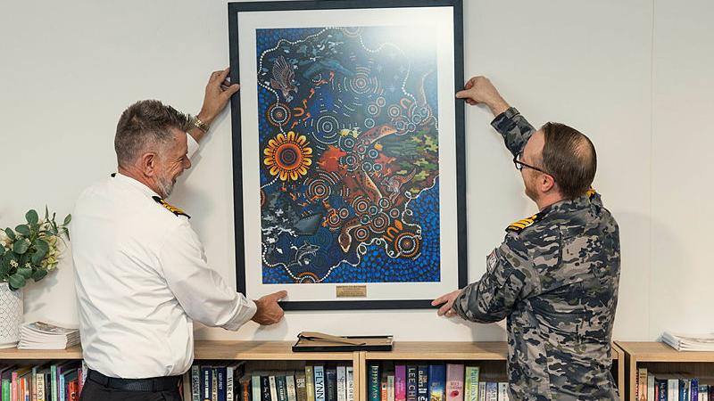 Navy Indigenous Adviser, Commander Michael Henry, and Commanding Officer HMAS Harman, Commander Dean Thompson, unveil and hang a print of Caina Putut, Ilya, Wartanganha by Chern'ee Sutton at a NAIDOC Week event.