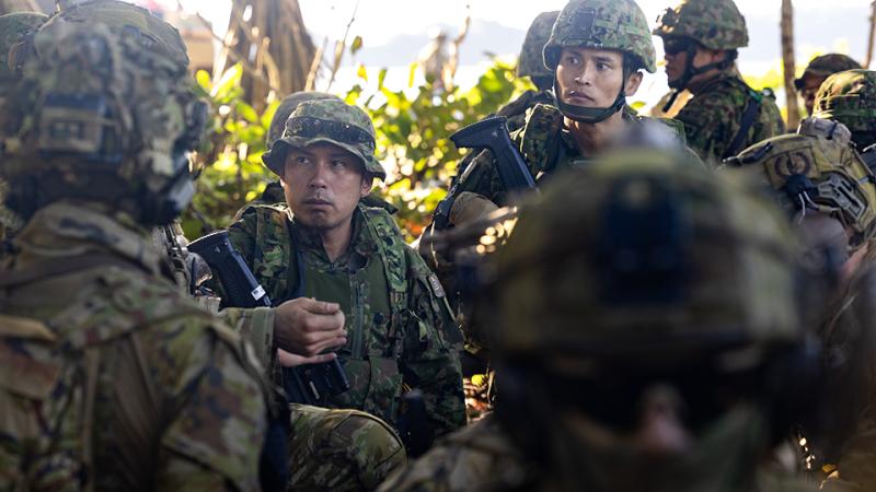 A group of soldiers from the Japan Ground Self-Defense Force discuss plans surrounded by foliage on the beach. 