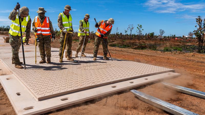 Soldiers in uniform and fluoro vests stand on a construction mat and prod it with poles.