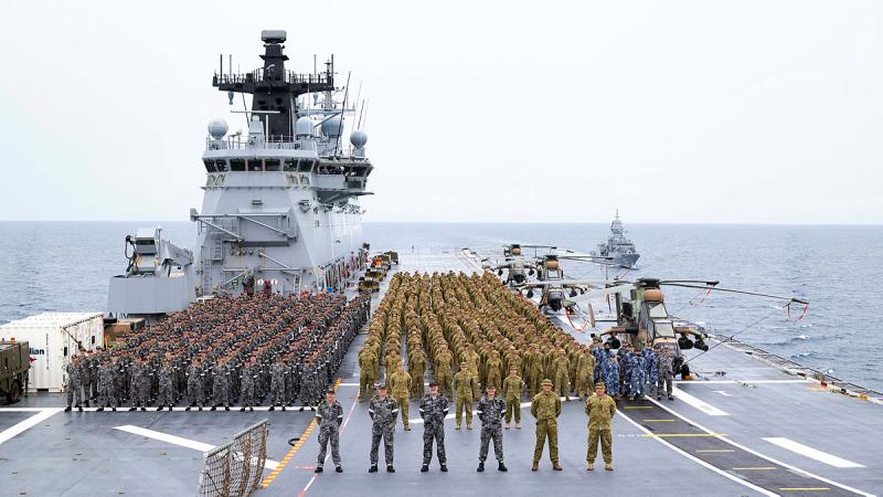 Navy, Army and Air Force personnel on parade on ship's flight deck with helicopters and military vehicles.