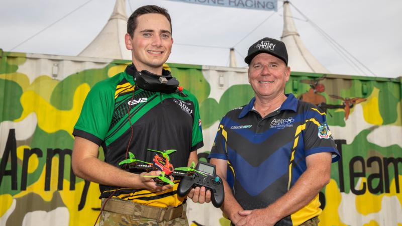 Two smiling men stand side by side, one holding a drone and its controls. In the background there is a green and yellow shipping container.