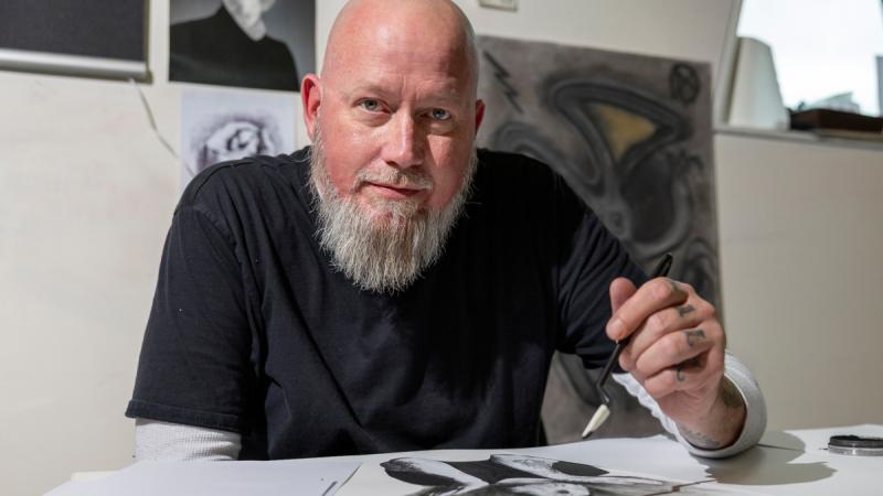 A bearded man holds a brush as he works on a black and white artwork.