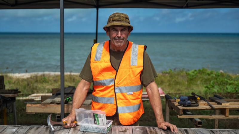 Sapper Stewart, holding a hammer and wearing a high-vis vest, stands in an open-air, waterfront workshop on a sunny day.