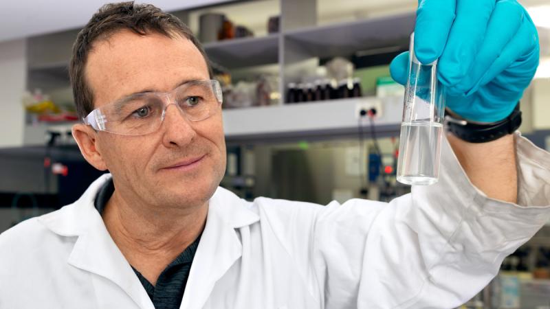 A man in a lab, wearing a lab coat, protective glasses and blue latex gloves, looks at a test tube he's holding.