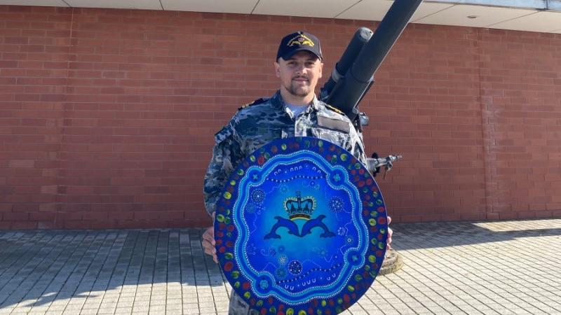 A Navy officer holds Indigenous artwork presented to HMAS Stirling, with a naval gun in the background
