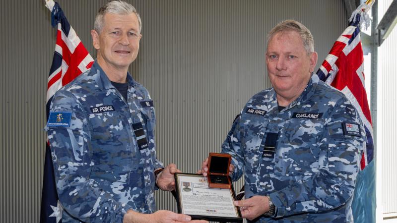 Two men in Air Force fatigues holding an award and standing in front of two Australian flags.