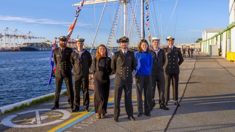 A group of people, several in Navy uniforms, standing in a 'V' formation in front of a tall ship.