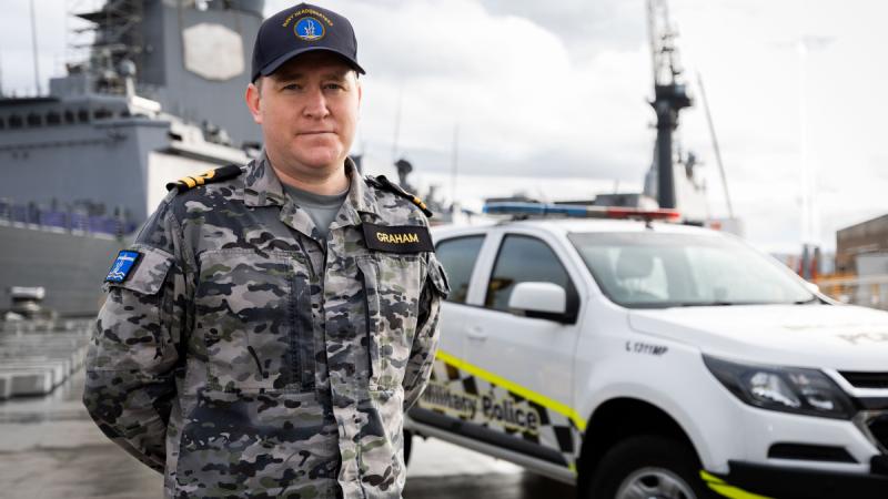 A man in Navy fatigues stands in front of a Military Police vehicle and a Navy ship.