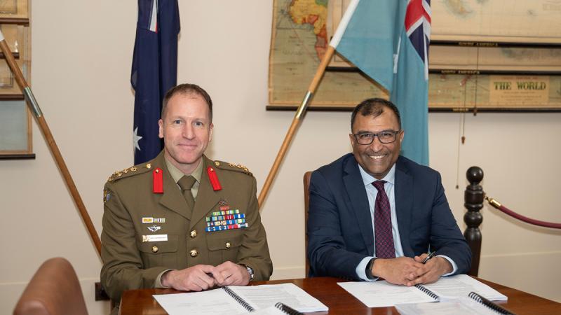 Two men, one in uniform, sit side by side at a document-laden table. Behind them are Australian flags and some maps.