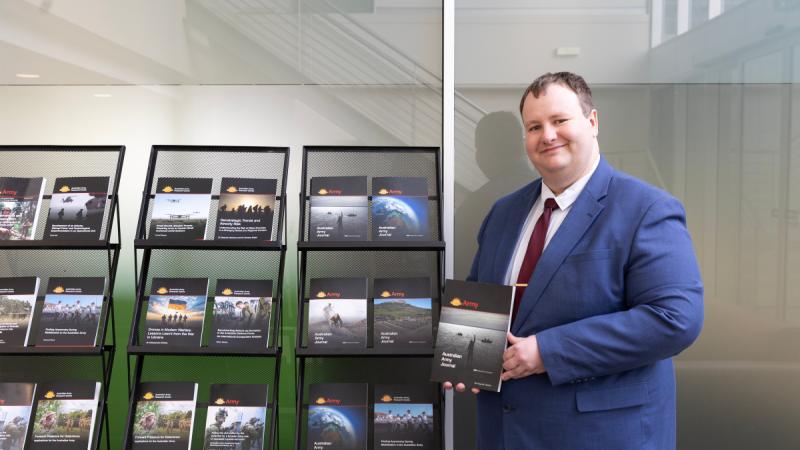 Dr Jordan Beavis holds a copy of the Australian Army Journal, in front of a display of past issues. 