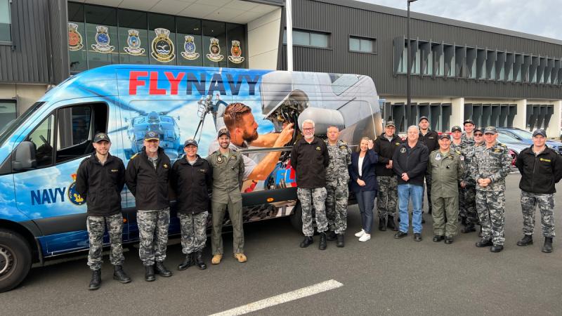 A group of approximately 15 people stand in front of a van covered in predominantly blue vinyl wrap and featuring the words 'Fly Navy'.