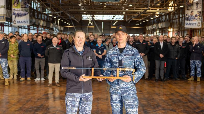 A woman and man in uniform holding a sword on a sword stand, with a large crowd of people standing behind them.