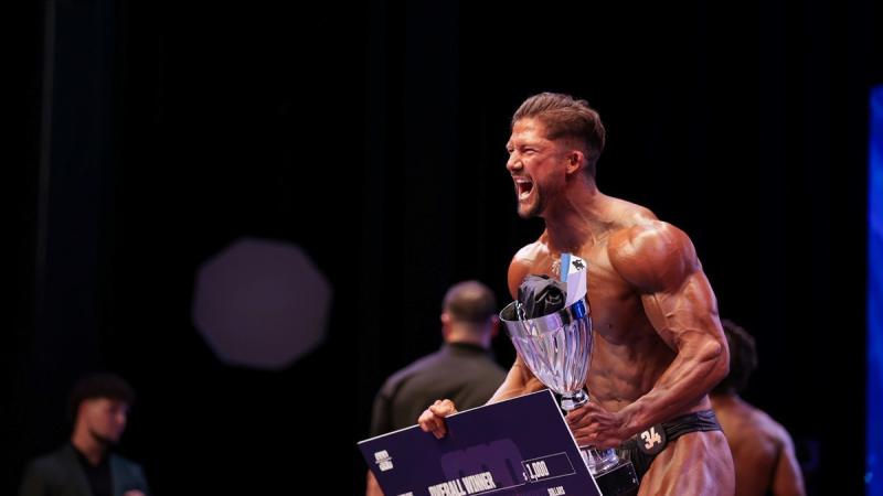 Corporal Gladman at a bodybuilding event, holding a novelty sized cheque and trophy, shouts in triumph.