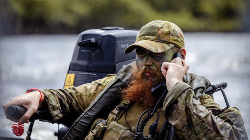 Alt caption: A soldier with a long, bright red beard listens to a communications radio while steering a small boat on a river.