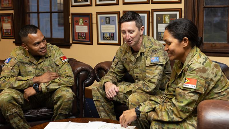 Three people in military uniform sit on leather armchairs, talking and looking at books on a coffee table.