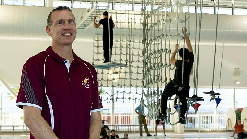 A man stands before a pool obstacle course with several other men on it.