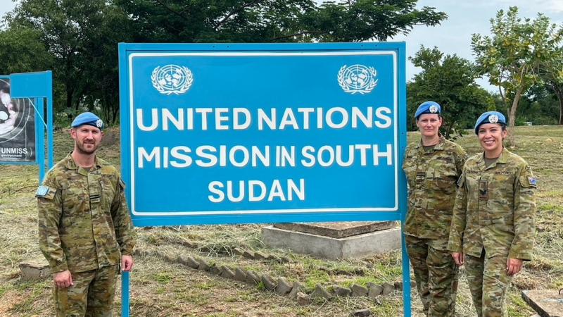 Three people in military uniform and light blue UN Peacekeeping berets stand in front of an UNMISS sign.