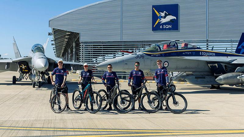 Five cyclists standing with their bikes in front of two aircraft.