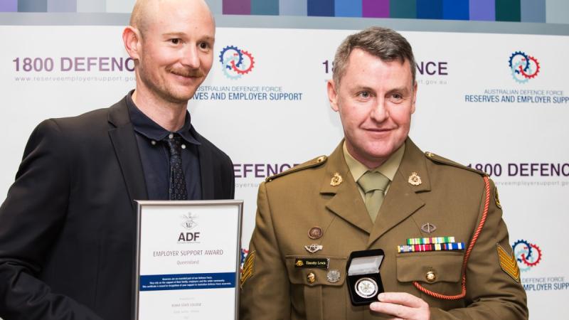 A soldier in Army uniform received an award on behalf of his civilian employer from a man in black suit, shirt and tie.