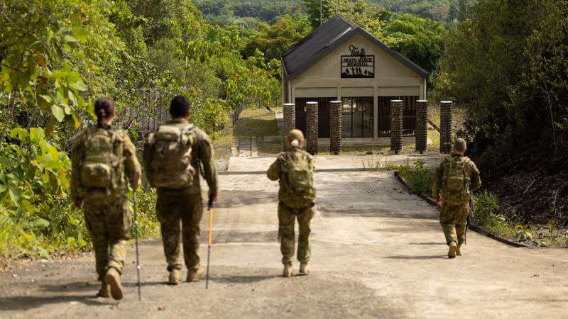 Four soldiers walking on a wide dirt road in a jungle setting towards a building bearing the sign ‘Death March Memorial’.