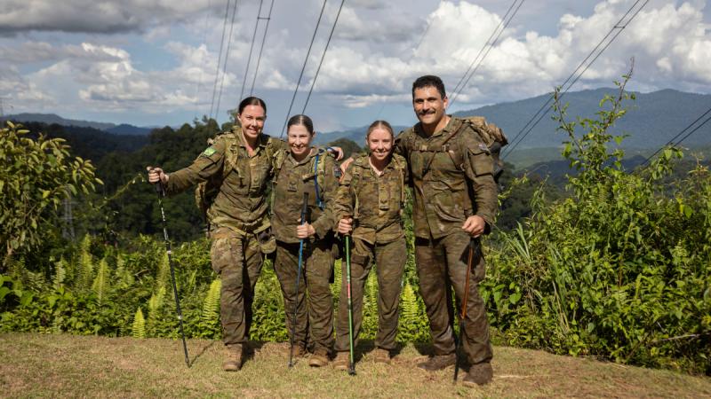 Four smiling people in Army fatigues hold hiking sticks as they stand at a point overlooking dense jungle.