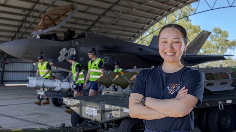 A young female technician stands in a hangar alongside other technicians working on a fighter jet.