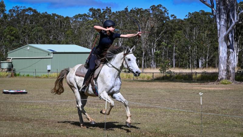 A woman riding on horseback fires and arrow from a bow.