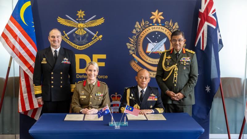 : Four smiling people wearing formal military uniforms behind a table, standing in between the Malaysian and Australian flags.