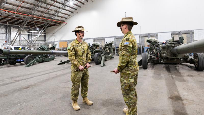 Two Army soldiers talk with weapons and equipment in the background.
