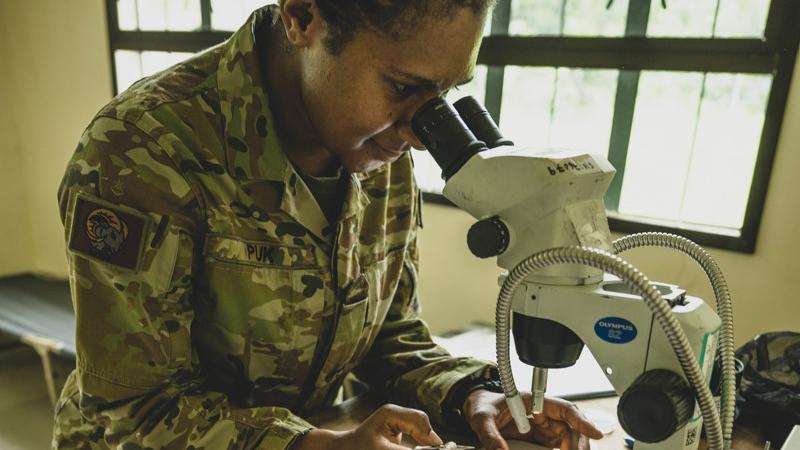 A woman in military fatigues looks into a microscope.
