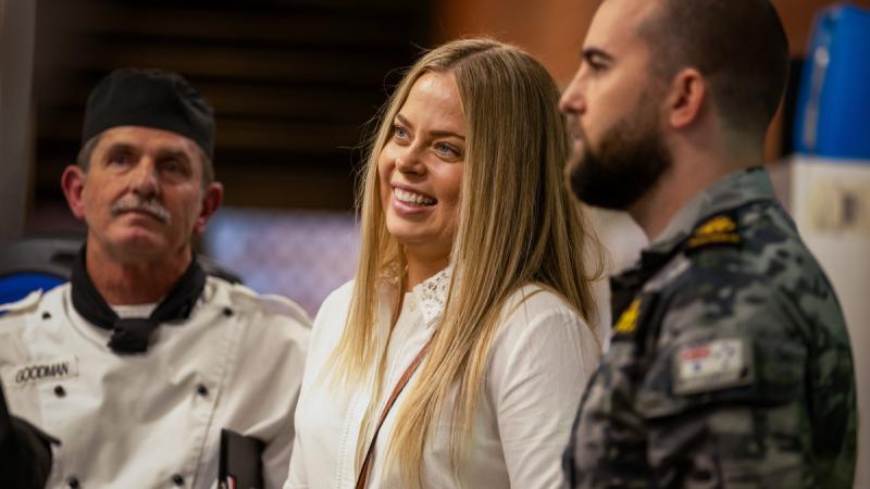 A woman with blonde hair stands between a chef and a Navy officer.