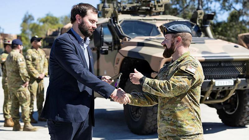 A man presents a key to another man in military uniform, in front of a military vehicle.