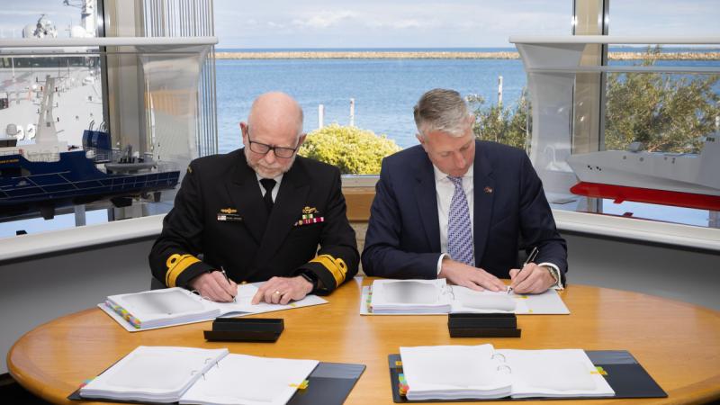 Two men signing some documents with a view of a harbour through the window behind them.