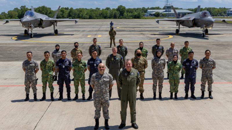 About 20 people standing in straight rows on a tarmac with hands clasped behind their backs, posing for a photo in front of two aircraft.