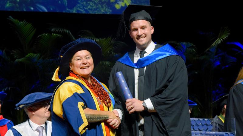 An academic in cap and gown awards a degree scroll to a tall man in university garb.