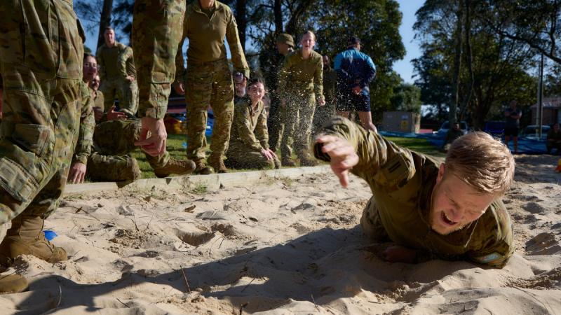 A man reaching out as he moves through a sandpit on his stomach. Spectators are shouting from the sidelines.