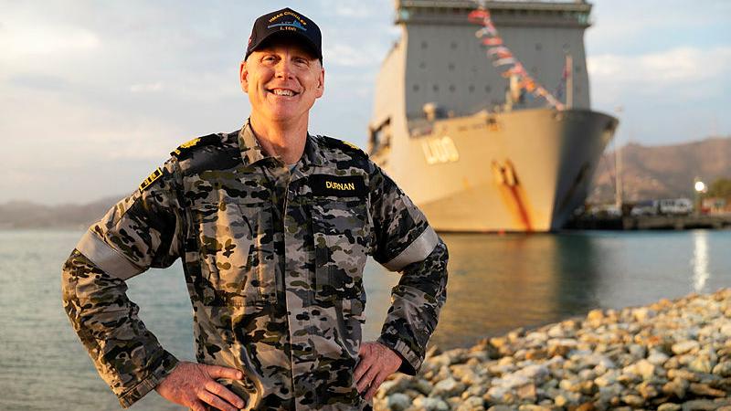 A man in uniform stands in front of a ship moored to a dock.
