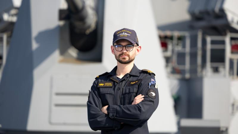 A man wearing a New Zealand Navy uniform stands on the forecast of a ship with his arms crossed.