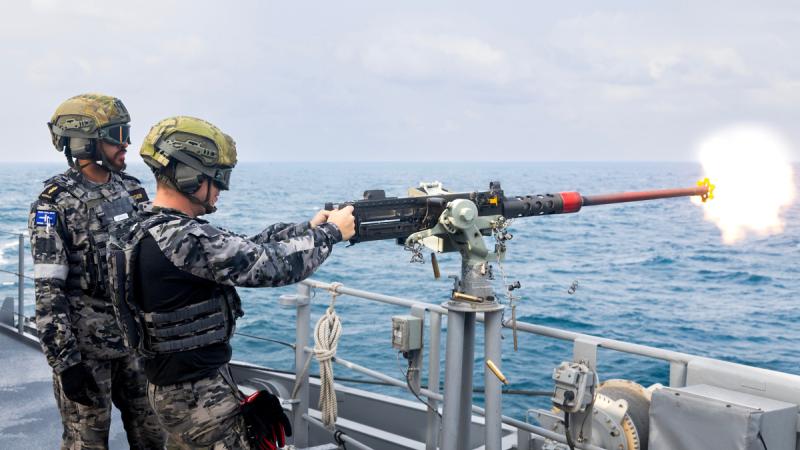 Two men in uniform. One shoots a stationary gun and the other watches.