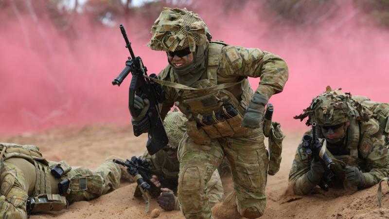 Soldiers running and crawling down a trench bearing weapons as pink smoke billows behind them.