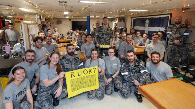 A group of people in a mess area holding a yellow sign that says 'R U OK?'.
