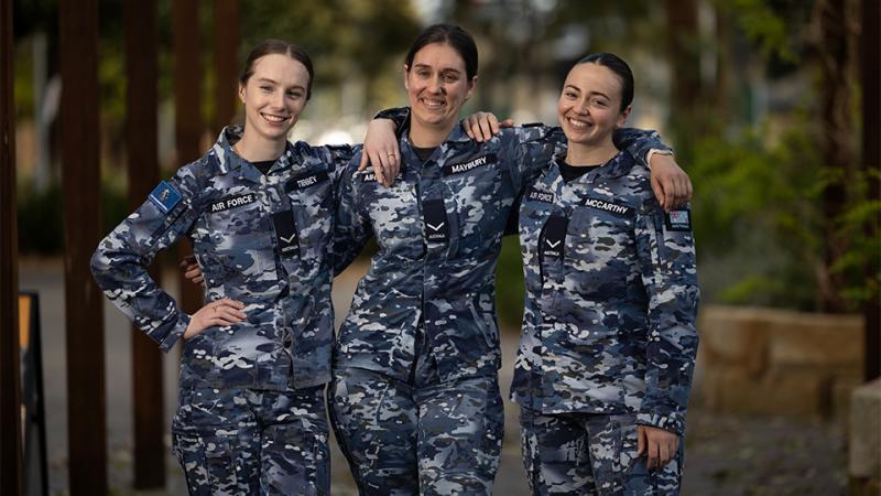 Three young woman in Air Force uniform stand with their arms around each other’s shoulders, smiling at the camera.