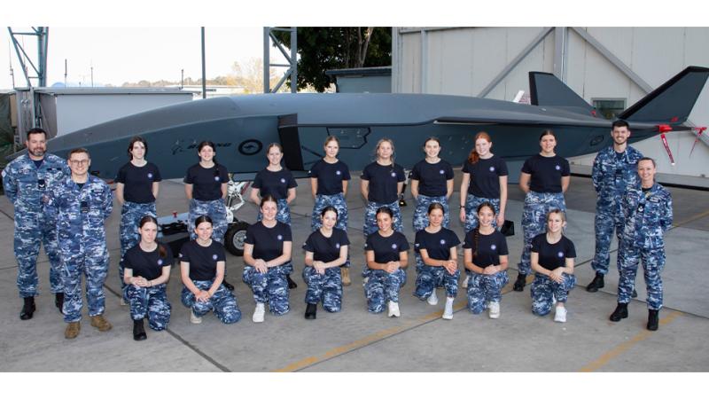 Two rows of people, mostly young women, with two RAAF personnel at each end, pose for a group shot in front of an unmanned aerial vehicle.