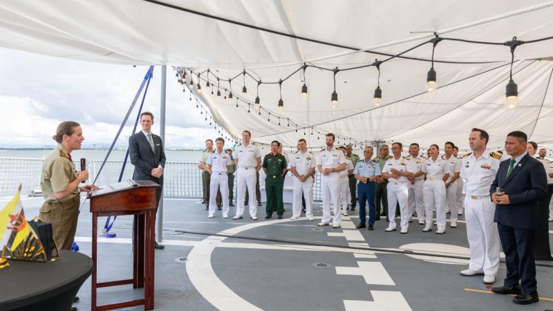 A woman in Army uniform giving a speech to an audience on the deck of a ship.