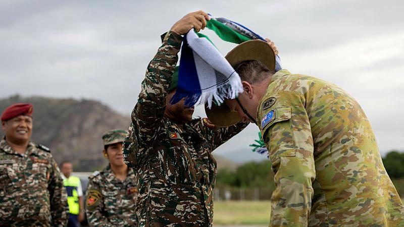 A man in military uniform places a scarf onto the shoulders of another man in military uniform.