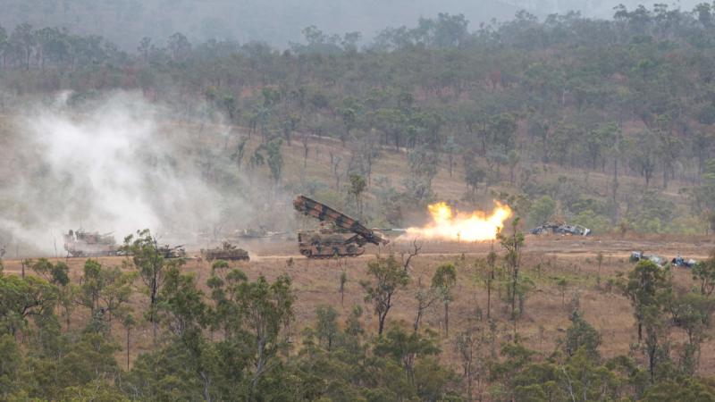 A long-distance photo of fire erupting from a tank, as several heavy armoured capability vehicles, including tanks and an assault bridge traverse scrubland.